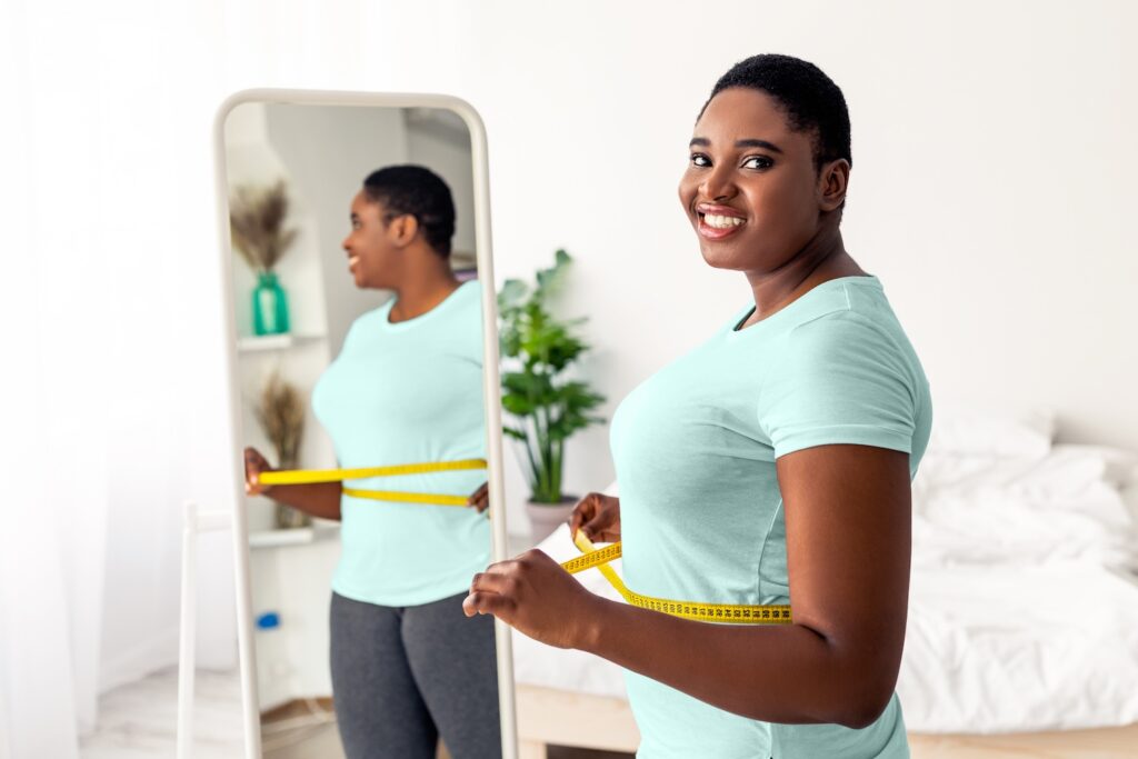 A young black woman measuring her waist with tape next to a mirror, after hearing about Retatrutide